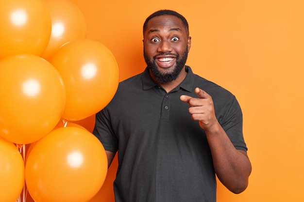 Positive bearded man with thick beard smiles positively points index finger directly at you holds bunch of inflated balloons wears black t shirt isolated over vivid orange wall