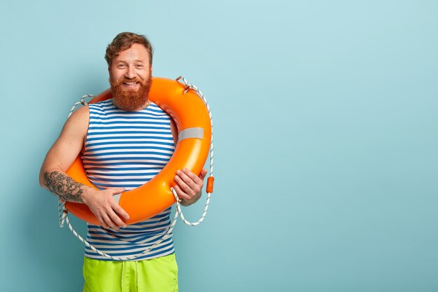 Позитивно бородатый рыжеволосый парень носит полосатый матросский жилет и несет оранжевый спасательный круг