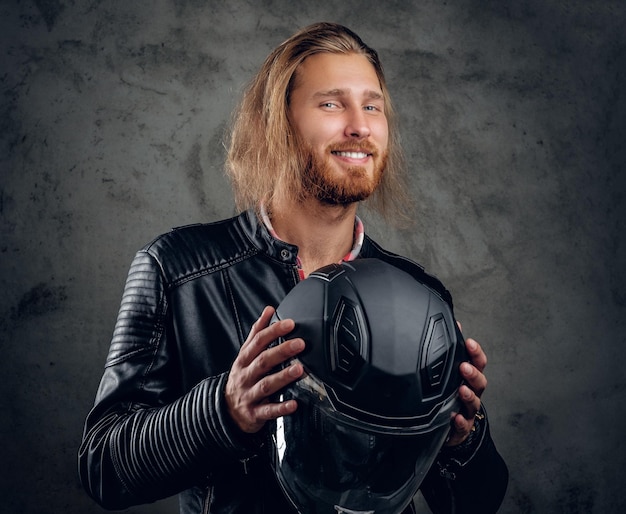 Positive bearded redhead male in leather jacket holds motorcycle helmet on grey background.