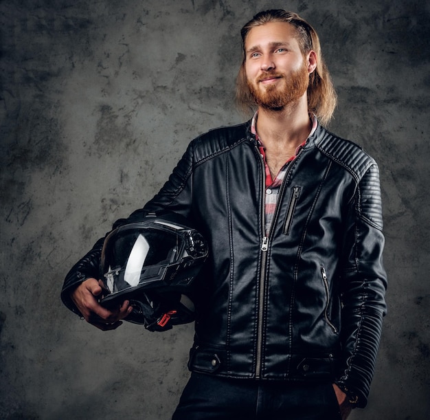 Positive bearded redhead male in leather jacket holds motorcycle helmet on grey background.