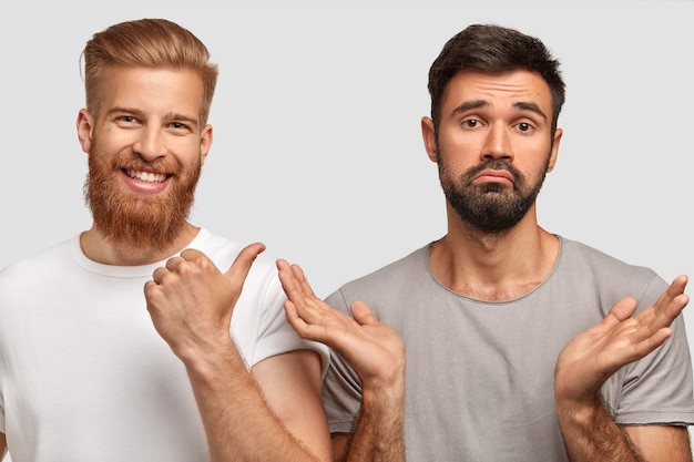 Positive bearded young red haired male with pleased expression indicates with thumb at his puzzled male friend who clasps hands and can`t make decision what to do, isolated over white wall