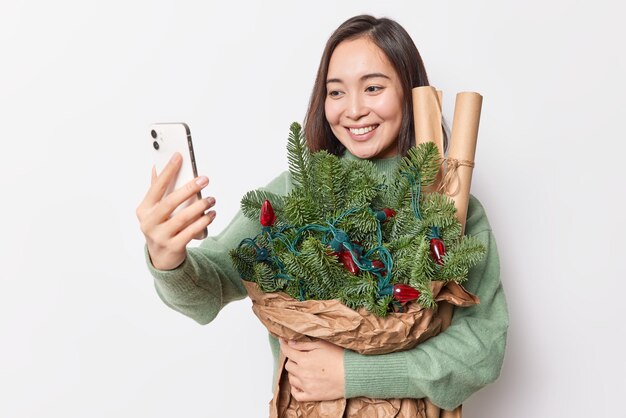 Позитивная красивая азиатка делает селфи на смартфоне, держит еловые ветки, завернутые в бумагу, украшенные ретро-гирляндами, счастливо улыбаясь на белом фоне. Подготовка к Рождеству