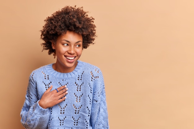 Positive beautiful female smiles gently and enjoys cheerful talk with someone concentrated aside happily dressed in knitted jumper isolated over beige wall copy space area