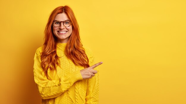 Positive beautiful ginger woman smiles broadly points right on blank space recommends good sale offer dressed in knitted sweater.