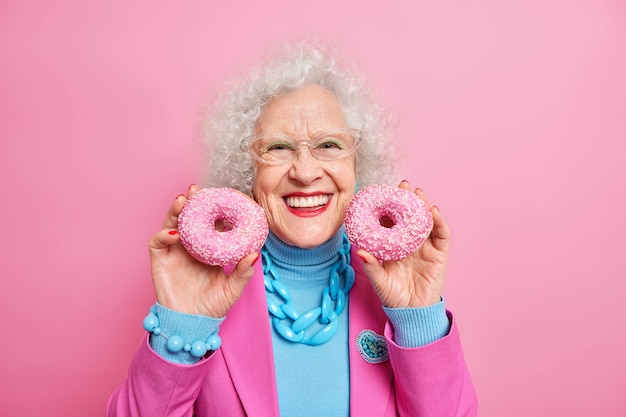 Positive beautiful wrinkled elederly European woman holds two delicious glazed doughnuts smiles broadly has good mood applies makeup fashionable clothes and jewelry