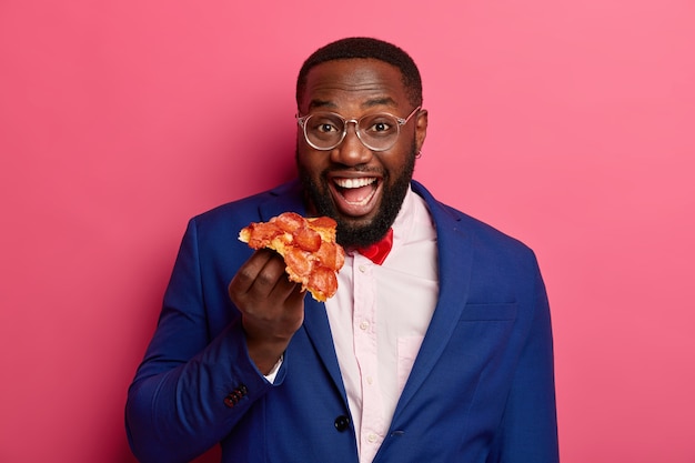 Positive black bearded man eats slice of pizza, wears formal clothing and transparent glasses, has good appetite, unhealthy snack