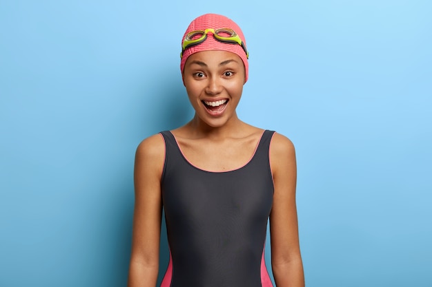 Positive black female swimming instructor dressed in swimsuit, swimcap and goggles