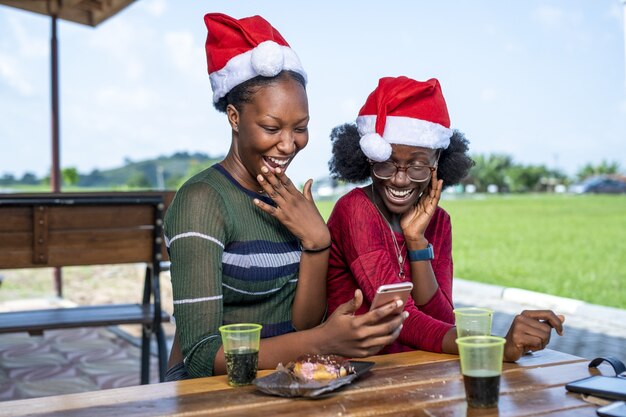 Positive Black friends in Santa hats having a video call while sitting outdoors at a picnic table