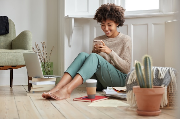Positive black lady enjoys coffee to go, holds smart phone in hands, reads text message in social networks, satisfied with modern technology and wifi connection, studies indoor at home, makes research
