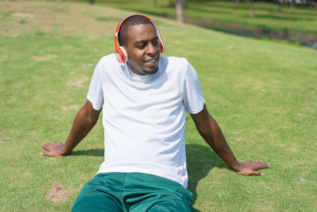 Positive black man relaxing in park and listening to music with wireless headphones.