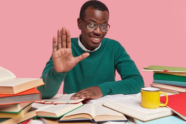 Positive black man shows palm, has optimistic look, greets with colleague or groupmate, has friendly look, sits at desktop, reads documentation