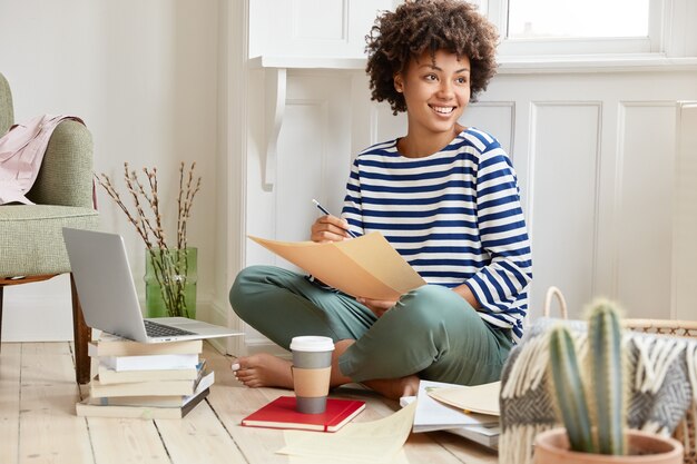 Позитивная темнокожая женщина сидит, скрестив ноги, одетая в полосатый матросский свитер, держит какие-то бумаги, занятые подготовкой отчета
