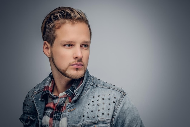 Positive blond bearded male dressed in a plaid shirt and denim jacket posing in the grey vignette background.