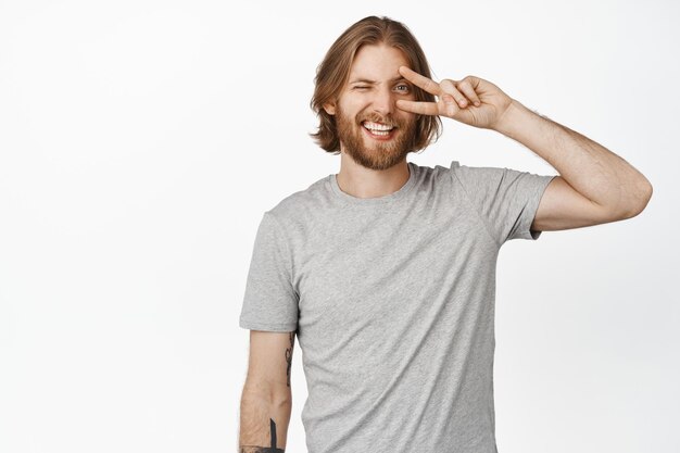 Positive blond bearded man showing peace v-sign near eye, kawaii gesture, winking and smiling happy, standing in grey tshirt against white background