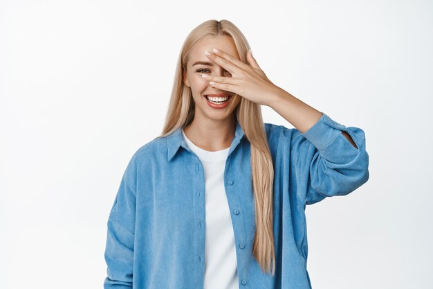 Positive blond girl laughing and smiling, peeking through fingers, open eyes, standing on white