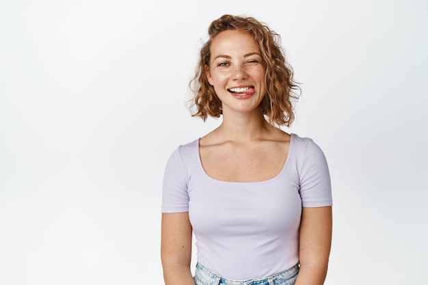 Positive blond girl shows tongue and winks at camera, makes happy and funny face, stands against white background