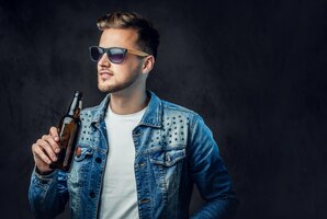 Positive blond male dressed in a denim jacket and sunglasses holds a bottle with craft beer.