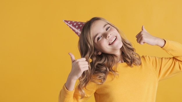Positive blond teenage girl in party hat looking happy keeping thumbs up on camera isolated on yellow background. Cheerful girl like her birthday party
