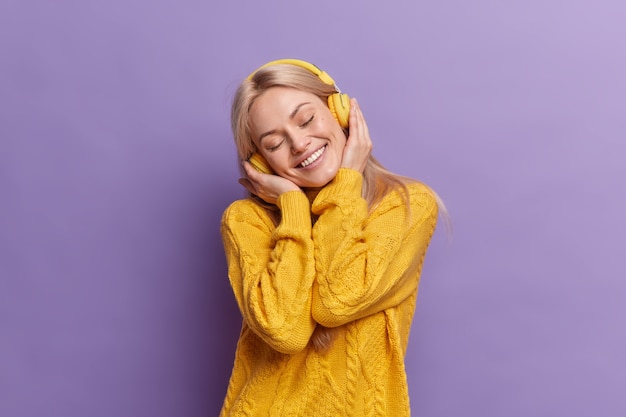 positive blonde European woman tilts head smiles broadly keeps eyes shut enjoys every bit of music wears wireless headphones dressed in yellow sweater 