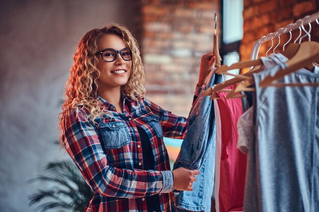 Positive blonde female in black eyeglasses chooses fashionable clothes on the coat rack.