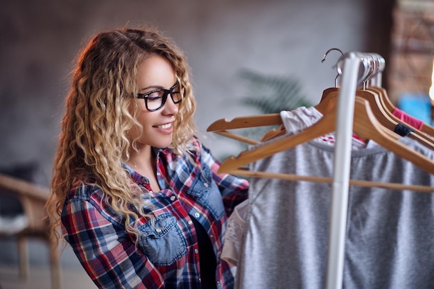 Positive blonde female in black eyeglasses chooses fashionable clothes on the coat rack.