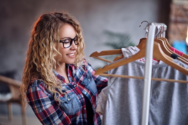 黒の眼鏡をかけたポジティブな金髪の女性は、コートラックでファッショナブルな服を選びます。