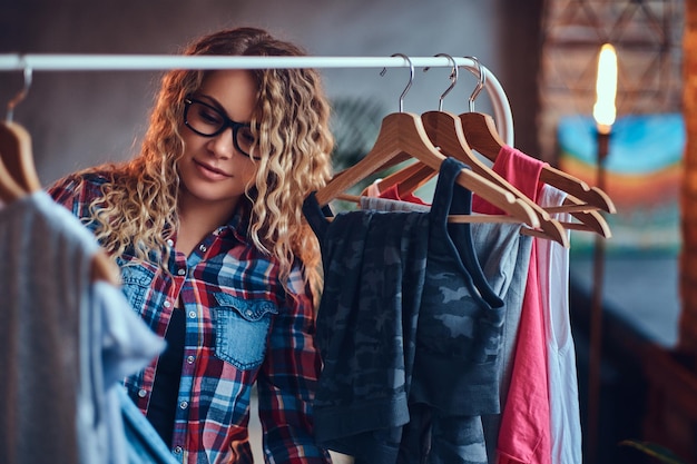 Positive blonde female in black eyeglasses chooses fashionable clothes on the coat rack.