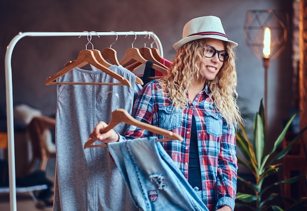 Positive blonde female in black eyeglasses chooses fashionable clothes on the coat rack.