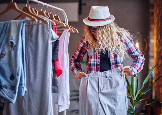 黒の眼鏡をかけたポジティブな金髪の女性は、コートラックでファッショナブルな服を選びます。