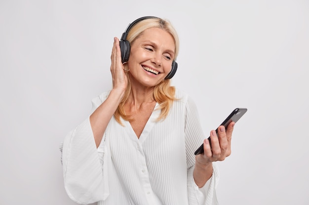 Positive blonde middle aged woman listens favorite music from playlist enjoys popular audio track in wireless headphones wears fashionable clothes isolated over white wall