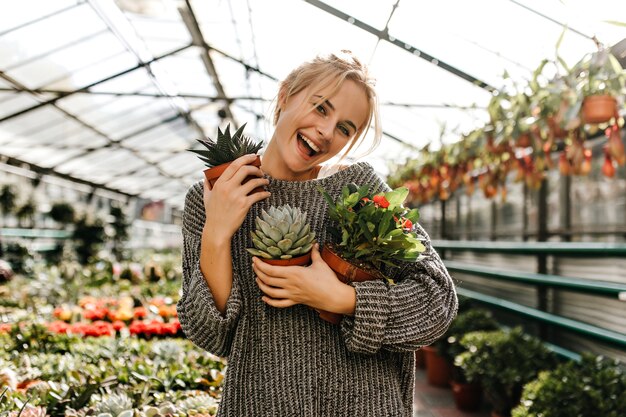 Positive blonde woman in knitted dress laughs happily, posing with different evergreens in greenhouse.