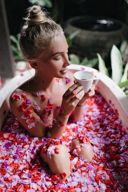 Positive blonde woman sitting in bath with flowers and holding cup. Portrait of romantic tanned woman drinking tea during spa.