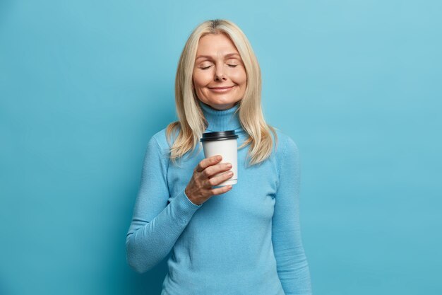 Positive blonde woman stands with eyes closed drinks takeaway coffee