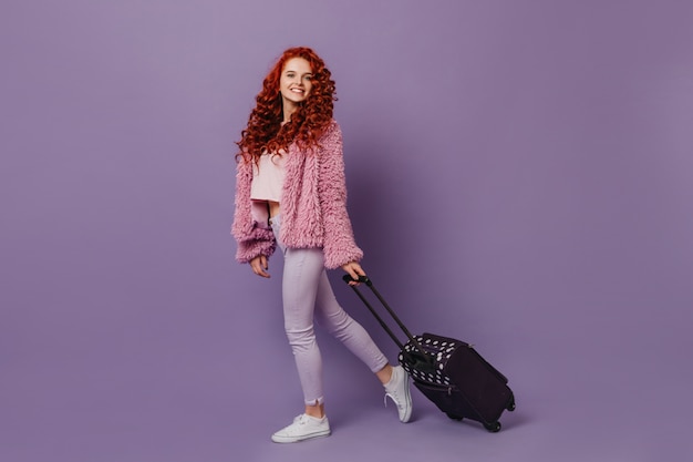 Позитивная голубоглазая девушка с рыжими кудрями, одетая в стильную шубу и белые брюки, несет чемодан.