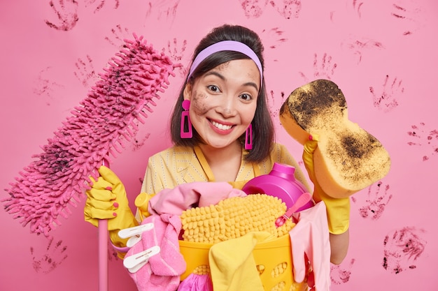 Positive brunette Asian housewife smiles pleasantly wears headband earrings rubber gloves holds dirty mop and sponge satisfied with results of house cleaning