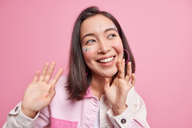 Positive brunette Asian woman smiles broadly has perfect white teeth face decorated with shiny stones dreamy cheerful expression keeps hands up wears denim jacket isolated over pink wall