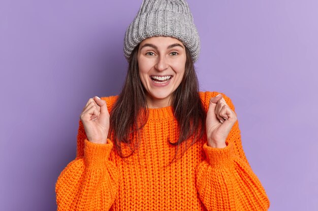 Positive brunette Caucasian woman raises clenched fists smiles positively awaits for something very good happen wears knitted sweater hat.