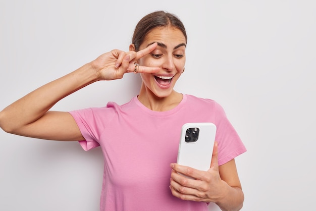 Positive brunette European woman makes peace gesture over eye holds mobile phone and makes selfie dresses in casual pink t shirt poses against white background People fun and technology concept