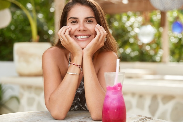 肯定的なブルネットの女性は、カフェテリアでゆっくり休んで、ピンクのスムージーを飲み、笑顔が広く、白い歯を見せ、夏休みに満足しています。