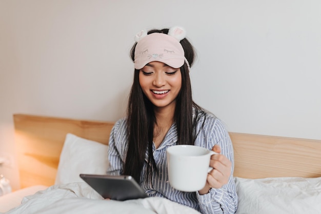 positive brunette girl reads and holds cup portrait of woman in sleeping clothes in bed