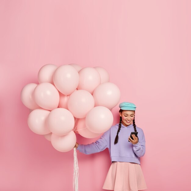 Positive brunette girl types messages on mobile phone, surfs internet, carries helium balloons