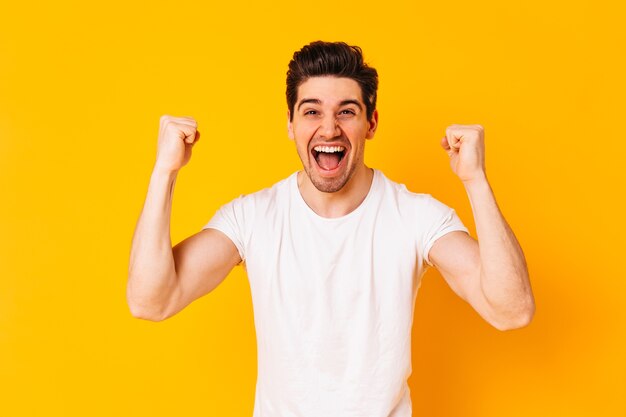 Positive brunette man rejoices in victory. Portrait of guy in white T-shirt on orange space.