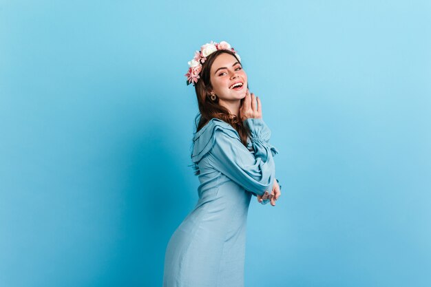 Позитивная брюнетка с цветами в вьющихся волосах смеется, позирует в атласном платье на синей стене.