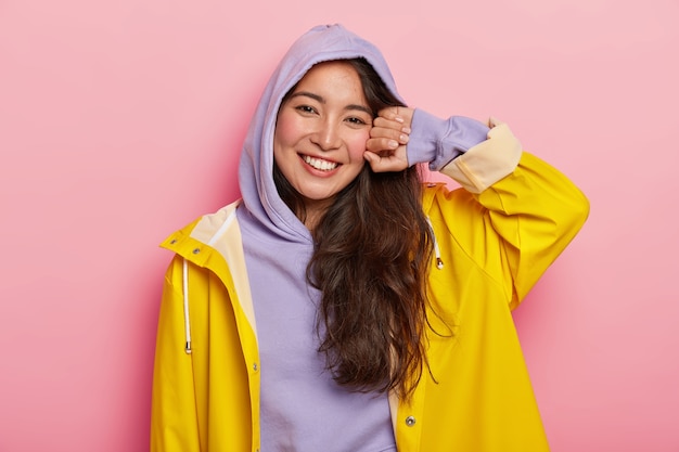 Positive brunette woman in casual hoody and raincoat, smiles pleasantly, being in good mood after outdoor walk with boyfriend