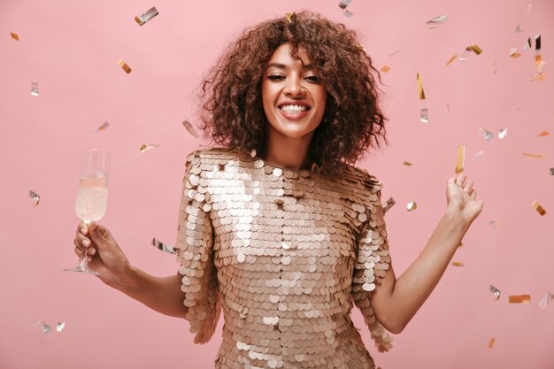 Positive brunette woman in modern beige dress smiling and posing with glass with wine and with confetti on pink wall..
