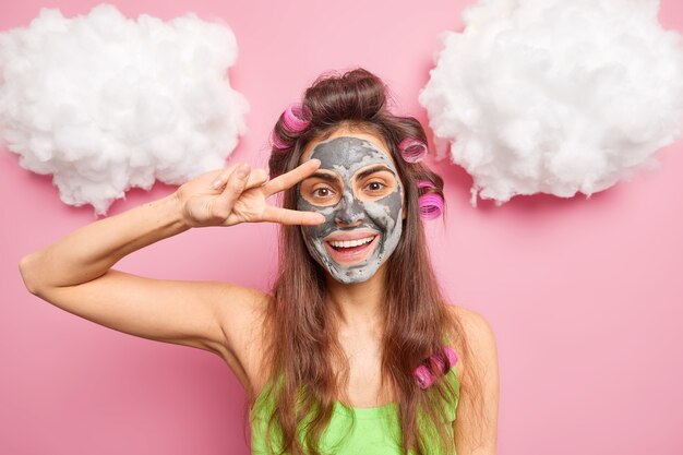 Positive brunette woman smiles gently makes peace gesture over eyes applies clay mask to reduce fine lines and blackheads makes perfect hairstyle at home poses indoor against pink wall