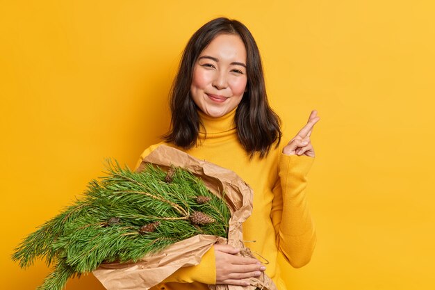 Positive brunette young woman carries spruce green bouquet for decorating house on new year makes wish and crosses fingers going to prepare christmas composition