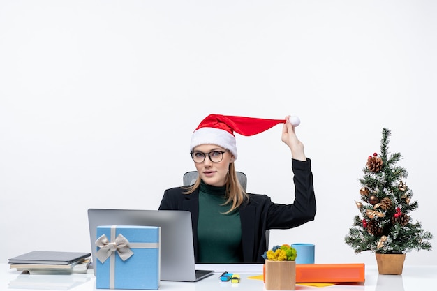 Positive business woman playing with a santa claus hat sitting at a table with a Christmas tree and a gift on it and checking her mails on white background