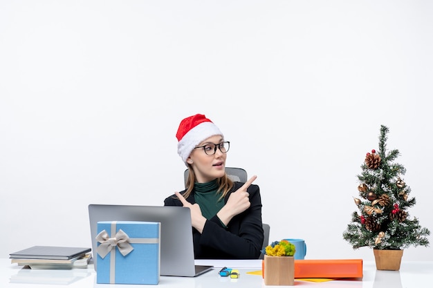 クリスマスツリーと白い背景の上の贈り物とテーブルに座っているサンタクロースの帽子を持つポジティブなビジネス女性