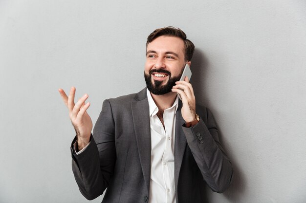 Позитивный деловой парень в формальных износа мобильного чата жесты при использовании мобильного телефона, изолированных на серый